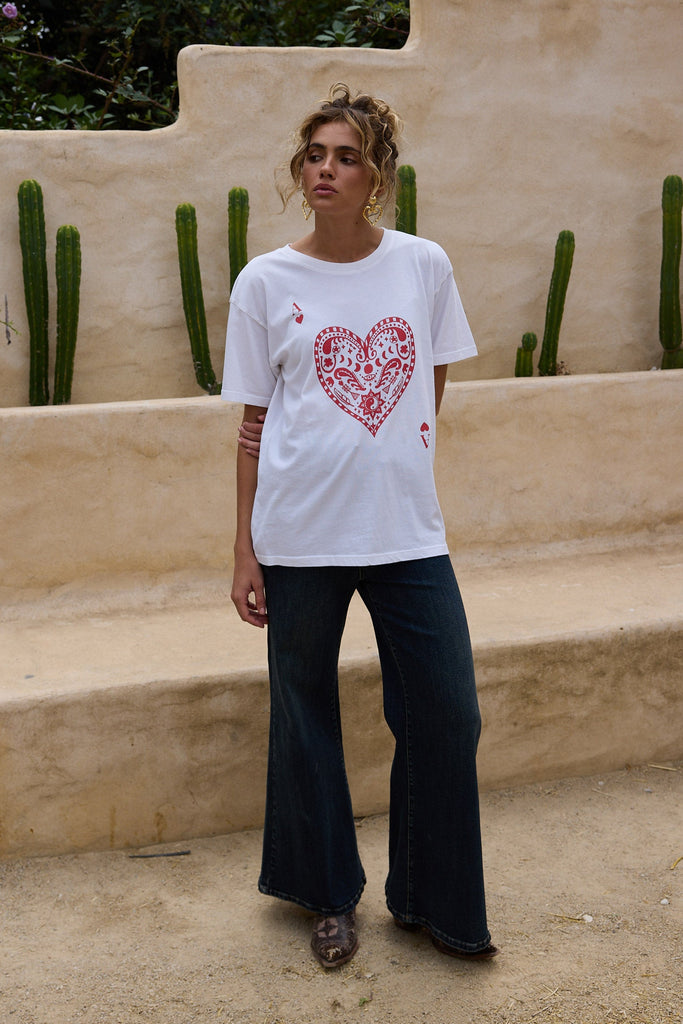 Person wearing a white t-shirt with a red heart design in an outdoor setting with cacti and a stone wall.