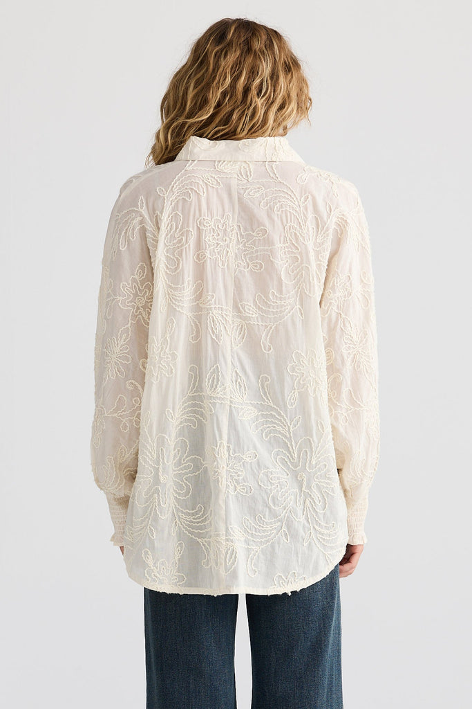 Person wearing a cream embroidered shirt and dark blue jeans on a white background