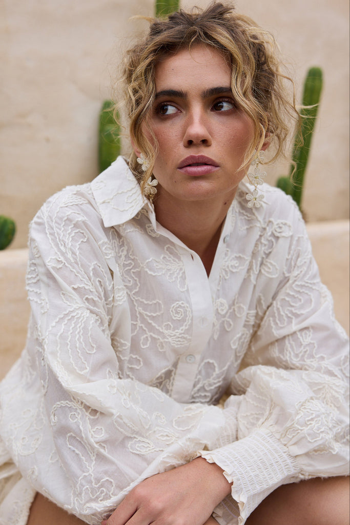 Woman wearing a cream embroidered shirt sitting against a beige wall with cacti.