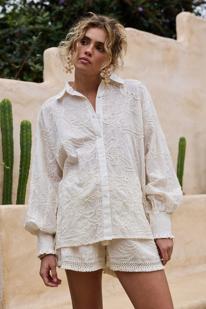 Woman wearing a natural embroidered outfit standing in front of a beige wall with cacti.