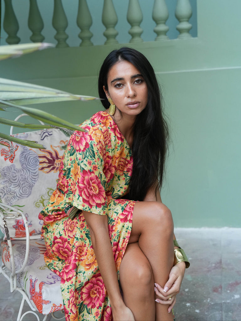 Woman in a colorful floral dress sitting outdoors with a light green wall and plant in the background.