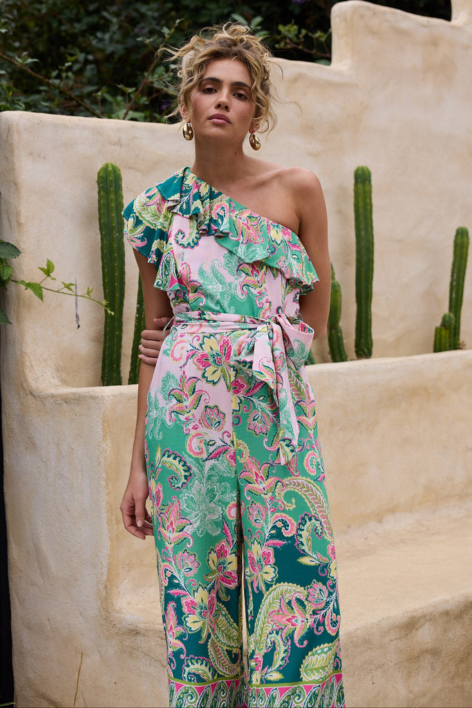 Woman wearing a colorful floral one-shoulder jumpsuit standing against a beige wall with cacti.