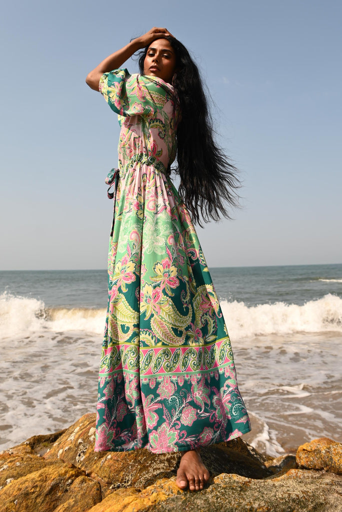 Woman in a colorful dress standing on a rock by the ocean with a clear blue sky.