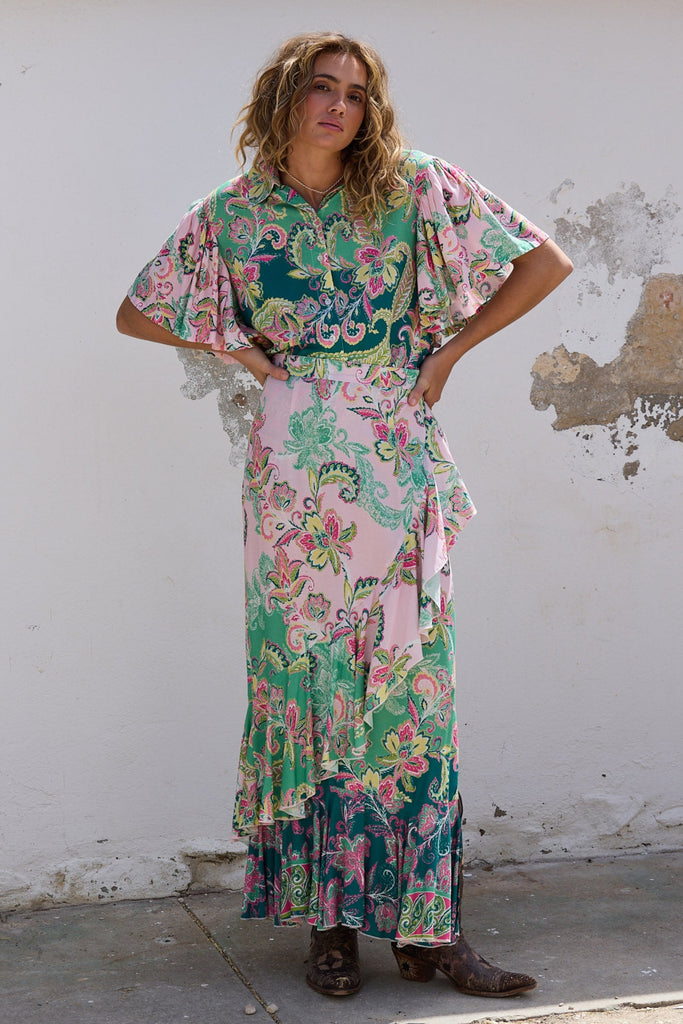 Woman in a floral dress standing against a white wall with a rustic building.