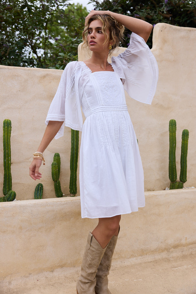 Woman in a white dress with sheer sleeves standing against a beige wall with cacti.