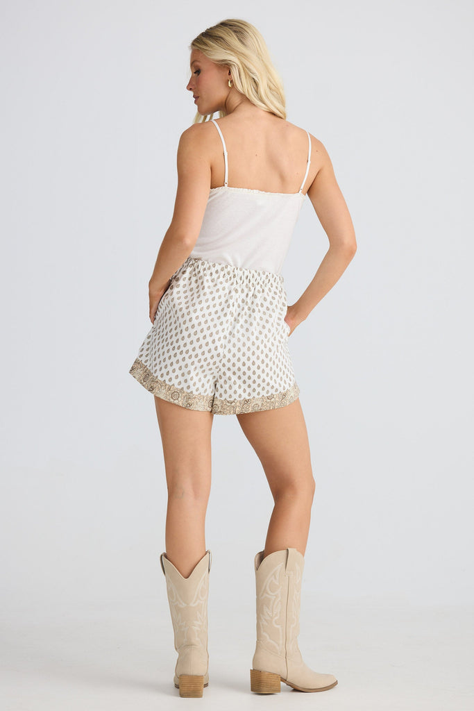 Woman wearing a white top and patterned shorts with beige cowboy boots on a plain background