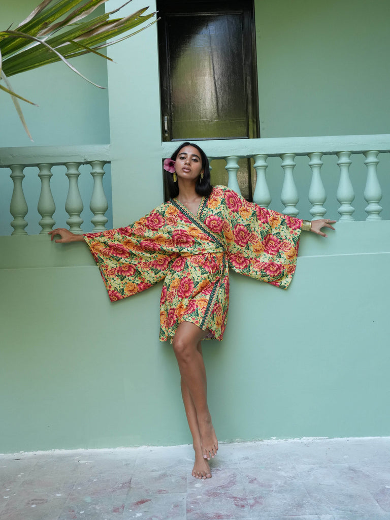 Woman in a colorful floral dress standing against a light green wall with a plant in the corner.