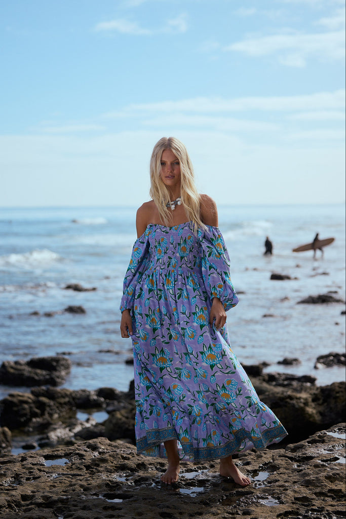 Woman in a floral dress standing on a rocky beach with ocean and sky in the background