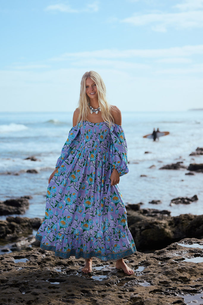 Woman in a floral dress standing on a rocky beach with ocean and sky in the background
