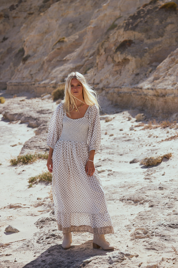 Woman in a long, light-colored dress standing on a rocky, desert-like landscape.