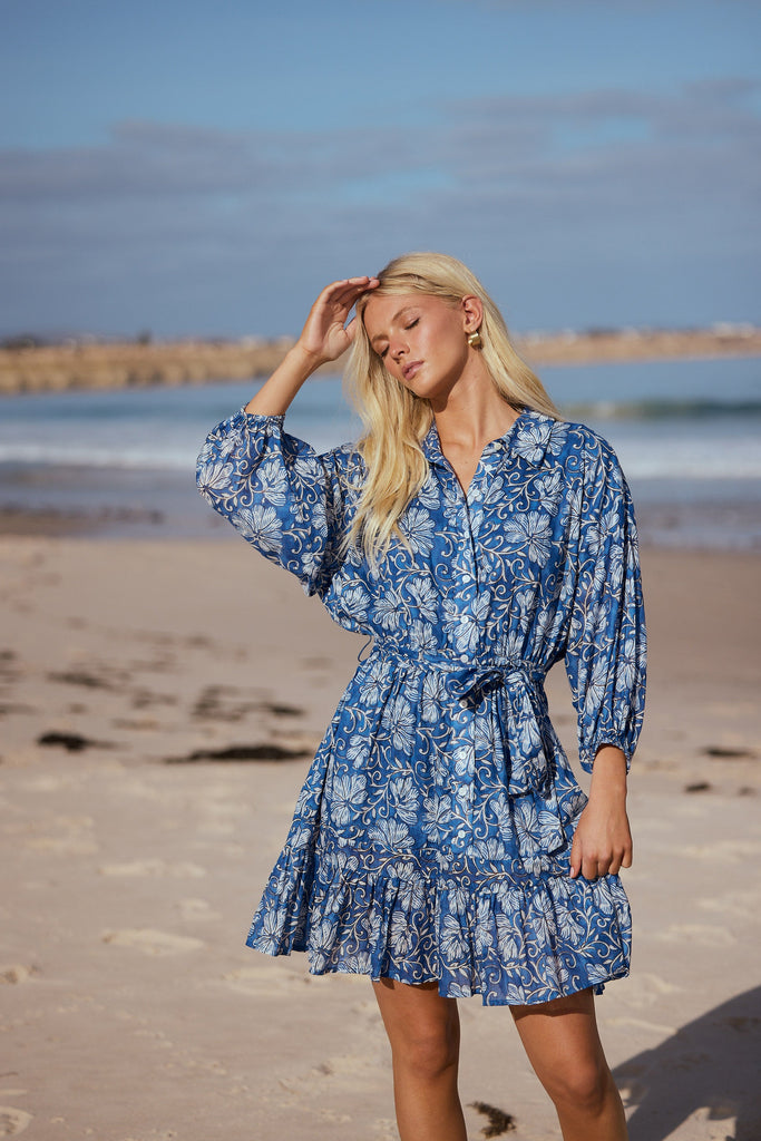 Woman in a blue floral dress standing on a beach with ocean in the background