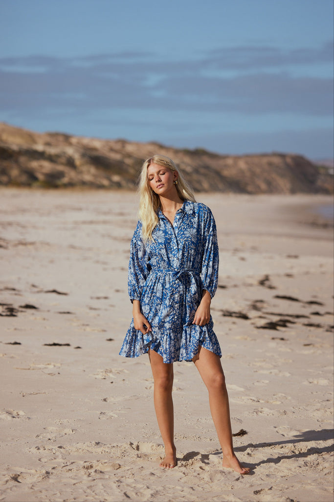 Woman in a blue dress standing on a sandy beach with a clear sky.