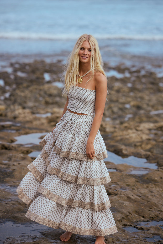 Woman in a white patterned skirt and top standing on a rocky beach.