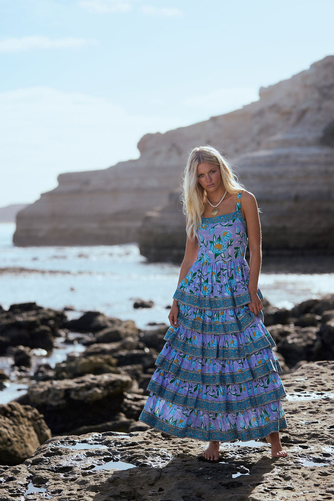Woman in a purple floral dress standing on a rocky beach with cliffs in the background
