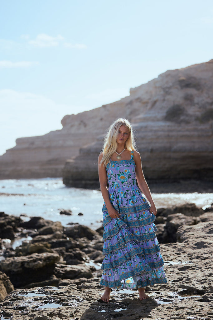 Woman in a purple patterned dress standing on a rocky beach with cliffs in the background.