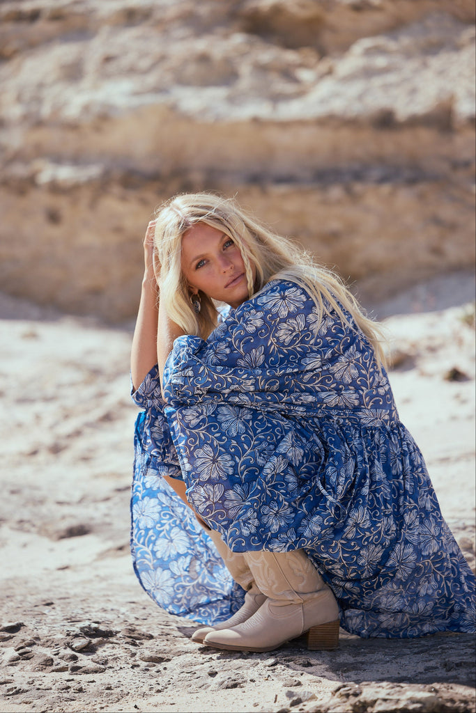 Woman in a blue floral dress sitting on a rocky beach.
