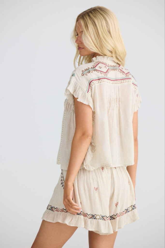 Woman wearing a off white embroidered top with tassels and matching shorts on plain white background
