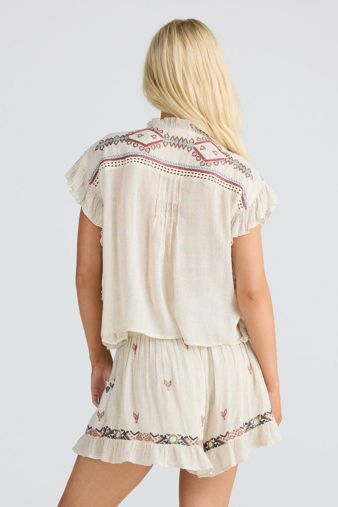 Woman wearing a off white embroidered top with tassels and matching shorts on plain white background