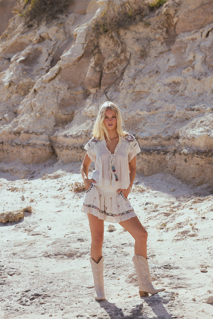 Woman wearing a off white embroidered top with tassels and matching shorts and beige cowboy boots standing on sandy beach