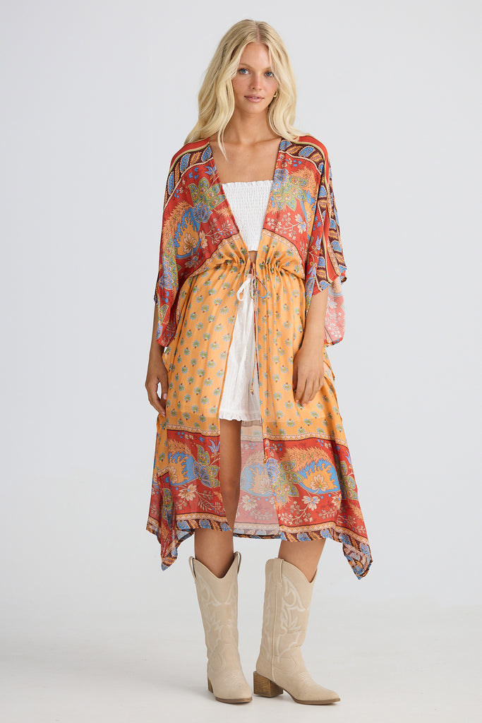 Woman wearing a colorful patterned cape with a white top and shorts and beige boots on a white background