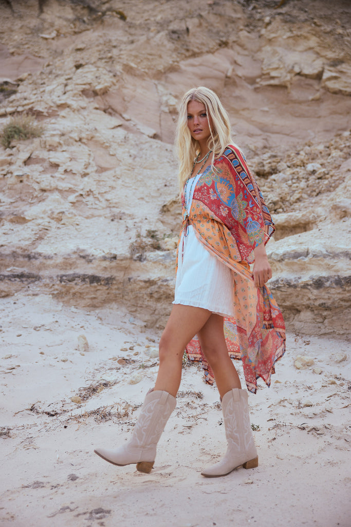 Woman in a colorful cape, white top and shorts and beige boots standing in a desert landscape
