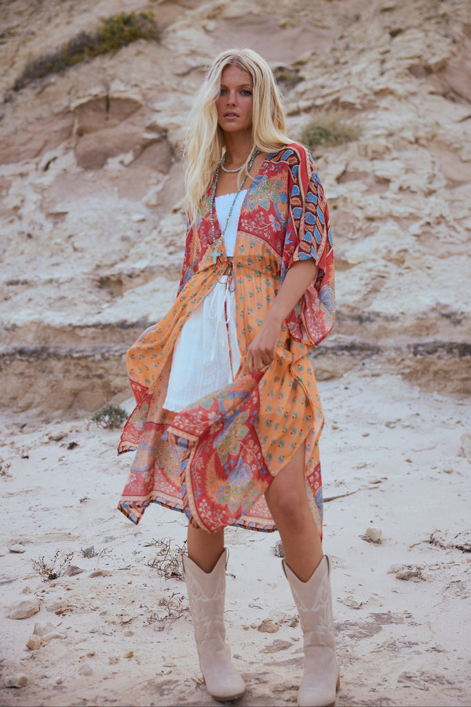 Woman in a colorful cape, white top and shorts and beige boots standing in a desert landscape