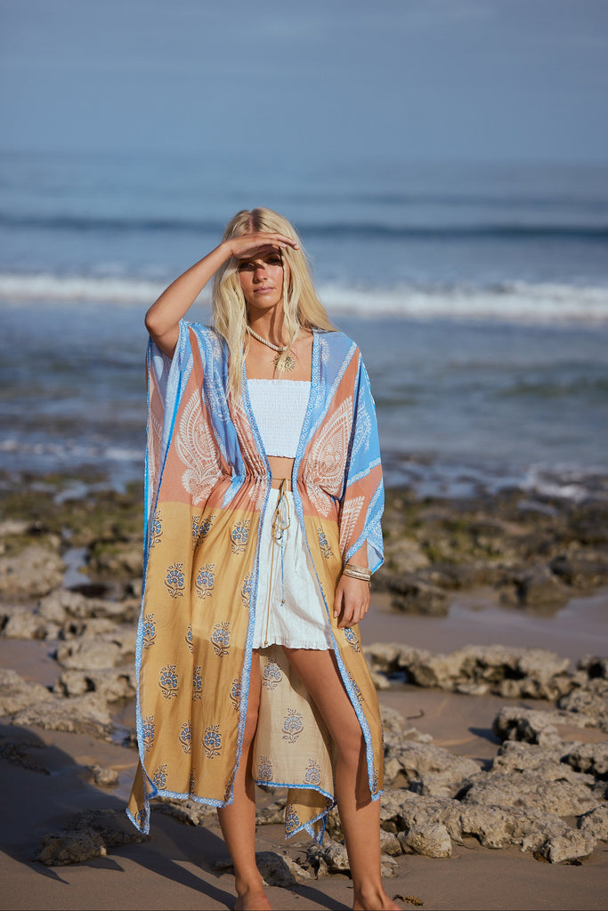 Woman in a long, patterned cape standing on a rocky beach with ocean waves in the background.