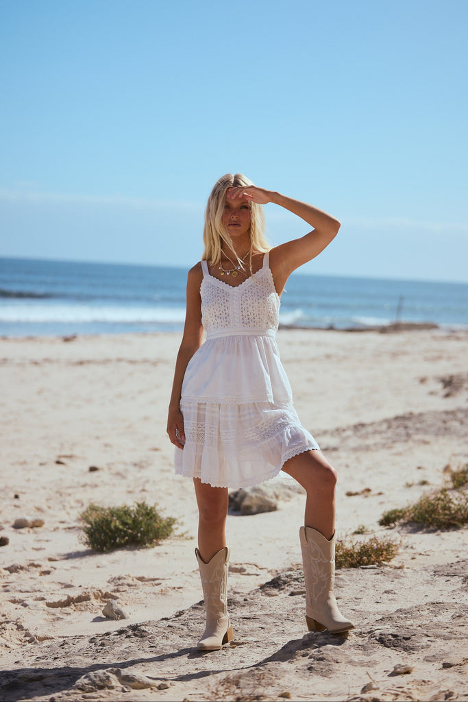Woman in a white singlet and skirt with boots standing on a sandy beach with ocean in the background