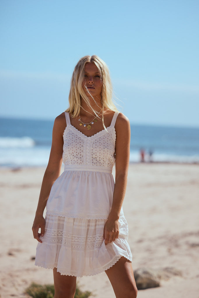 Woman in a white singlet and skirt standing on a beach with clear blue sky and ocean.