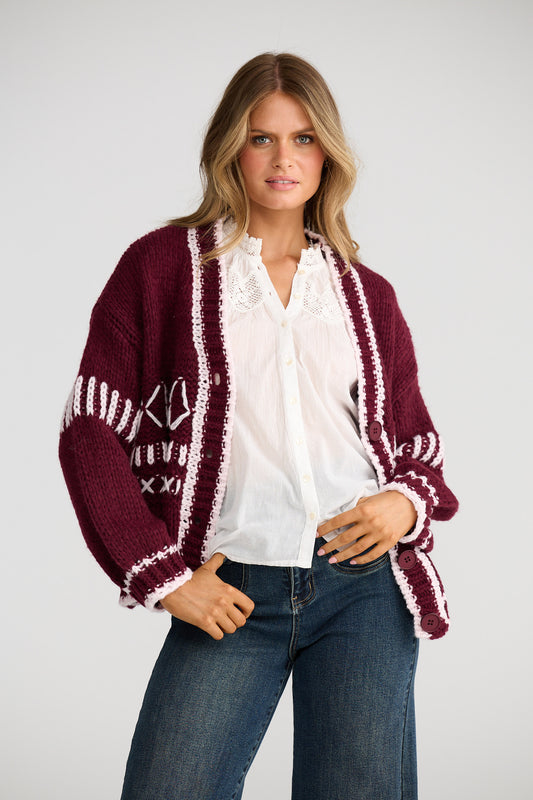 Woman wearing a burgundy cardigan with white patterns, white blouse, and blue jeans on a white background