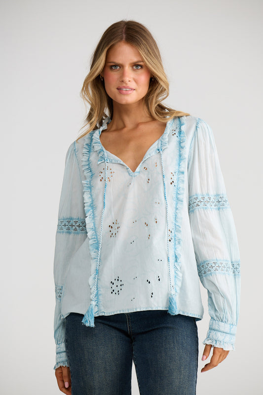 Woman wearing a light blue embroidered blouse over a white top with dark jeans on a plain background