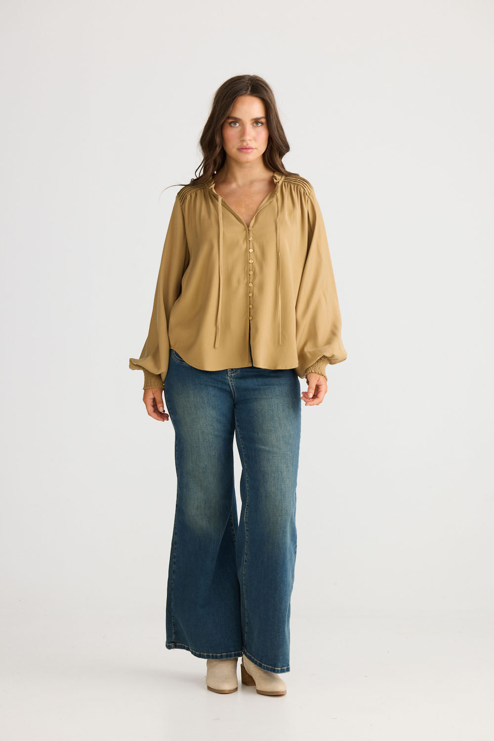 Model wearing Twilla Top in camel with round neckline and mini ruffle trim, side view on white background