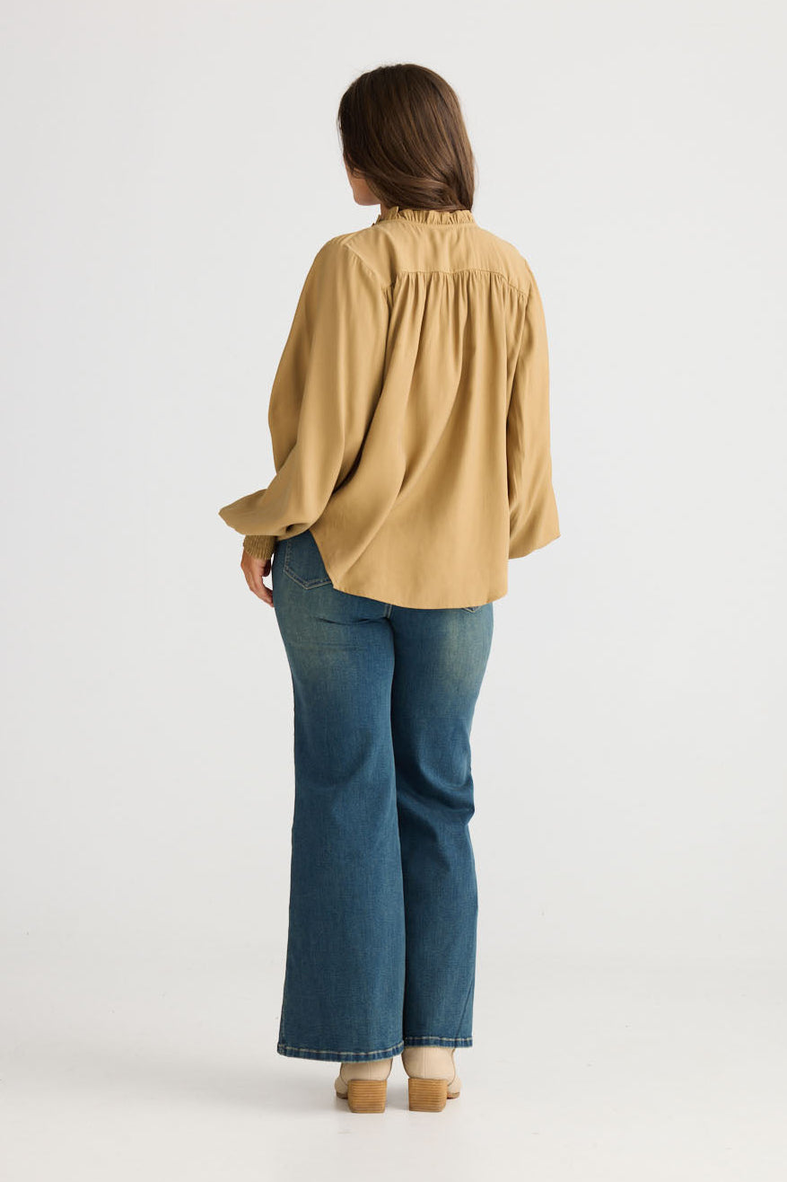 Model wearing Twilla Top in camel with round neckline and mini ruffle trim, back view on white background