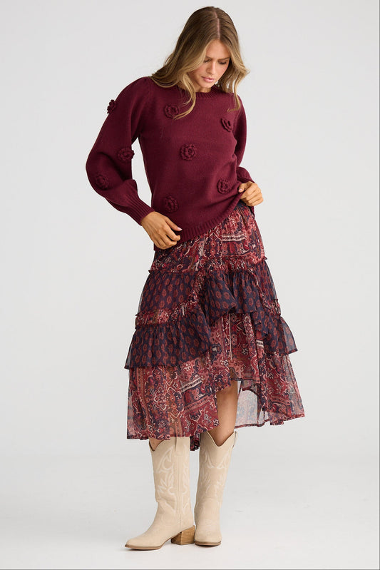 Woman wearing a burgundy sweater and patterned skirt with white boots on a light gray background