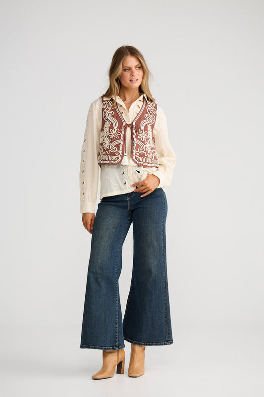 Woman wearing a patterned vest over a white shirt with blue jeans on a plain background
