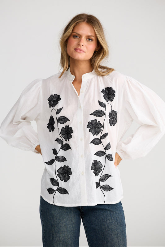 Woman wearing a white blouse with black floral embroidery on a plain background