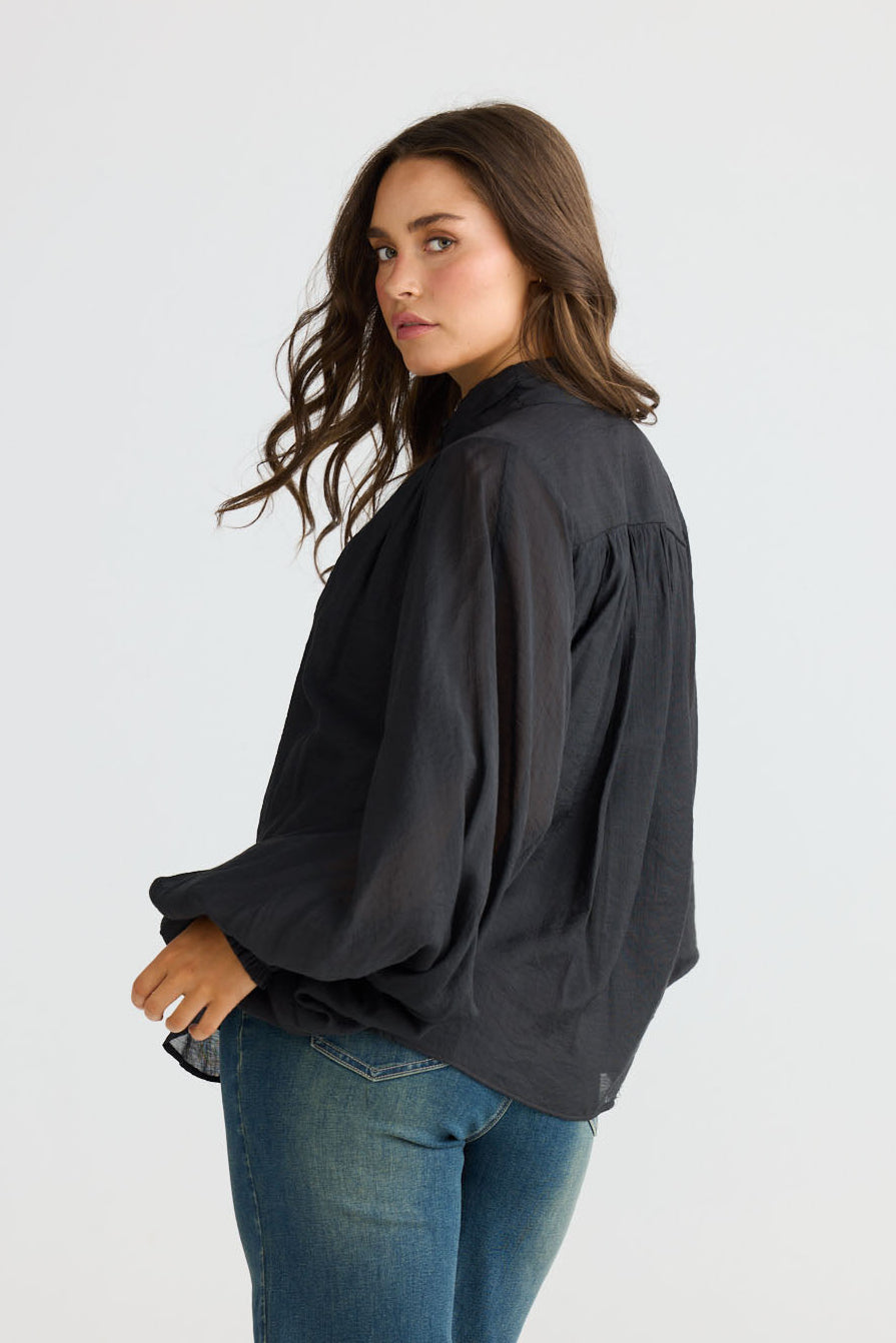 Model wearing Sylva Blouse in black with high collar with ruched details, back view on white background