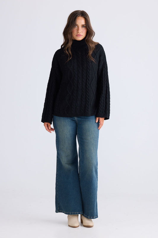 Model wearing Dixie Knit in black with roll-neck collar and cable knit design, front view on white background
