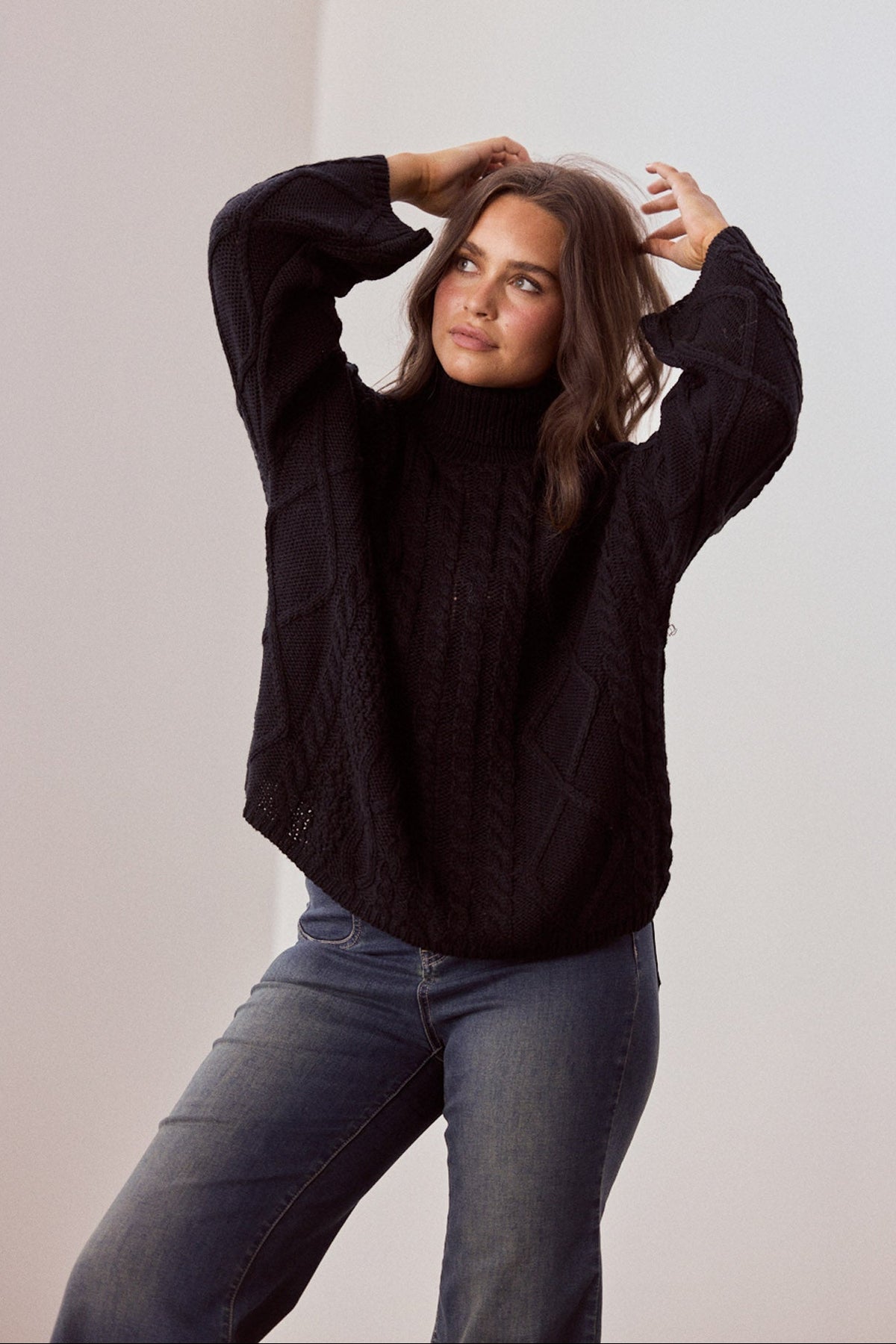 Model wearing Dixie Knit in black with roll-neck collar and cable knit design, front view on white background