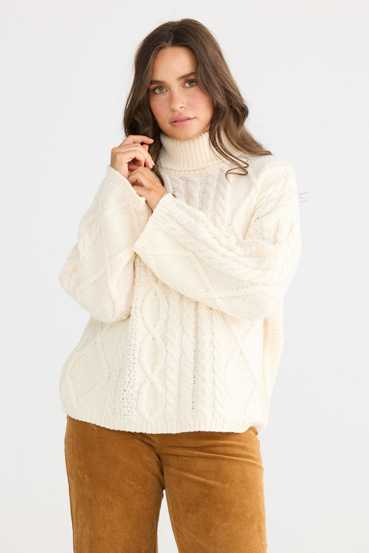 Model wearing Dixie Knit in cream with roll-neck collar and cable knit design, side view on white background