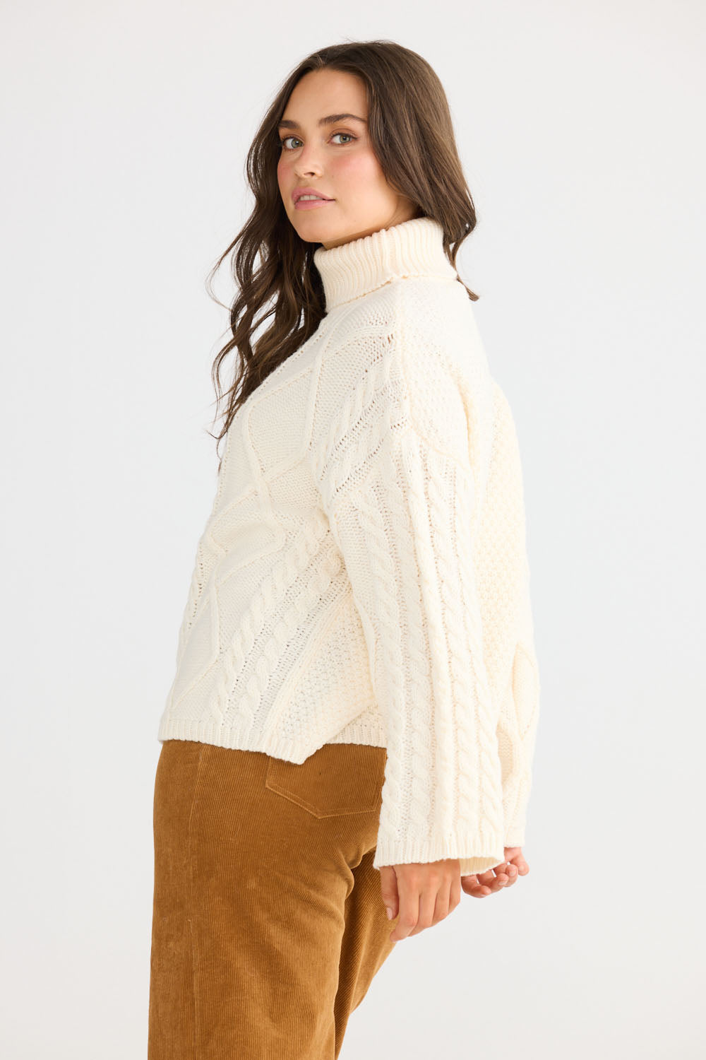 Model wearing Dixie Knit in cream with roll-neck collar and cable knit design, side view on white background
