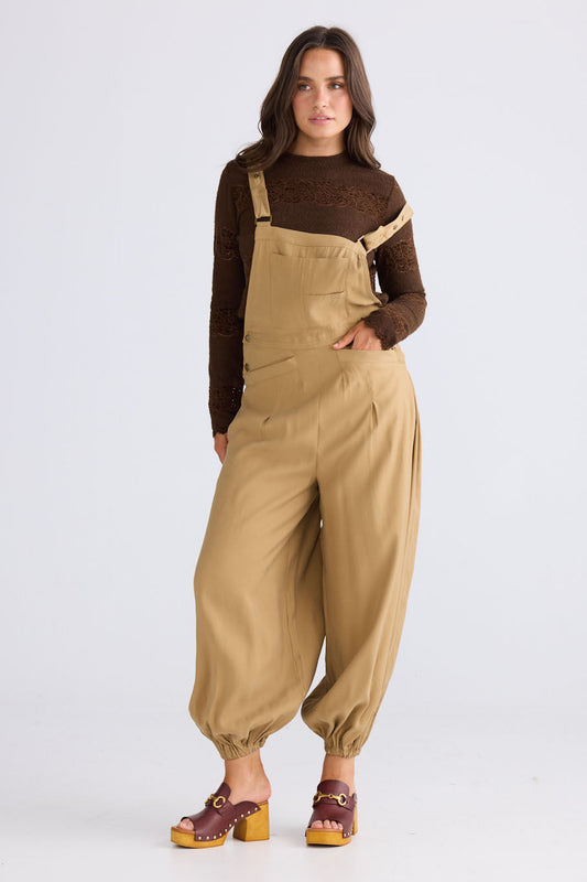 Model wearing Genie Overalls in camel with adjustable straps with antique brass button closure, back view on white background