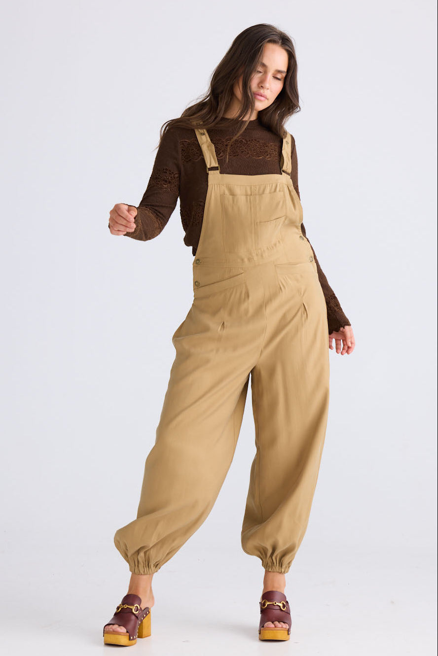 Model wearing Genie Overalls in camel with adjustable straps with antique brass button closure, side view on white background