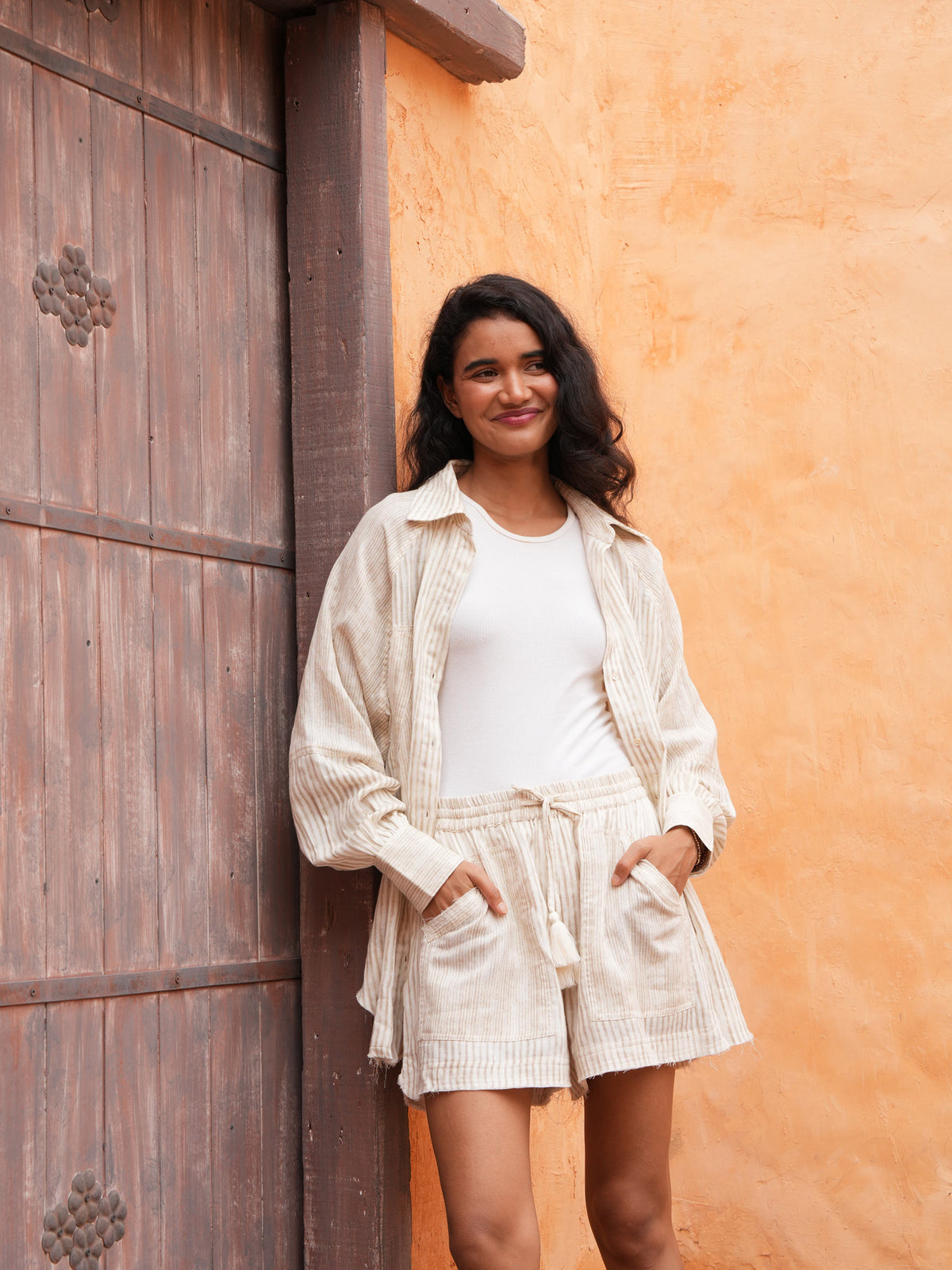Bodhi Shirt and Bodhi Short in natural stripe, standing at a weathered timber door.