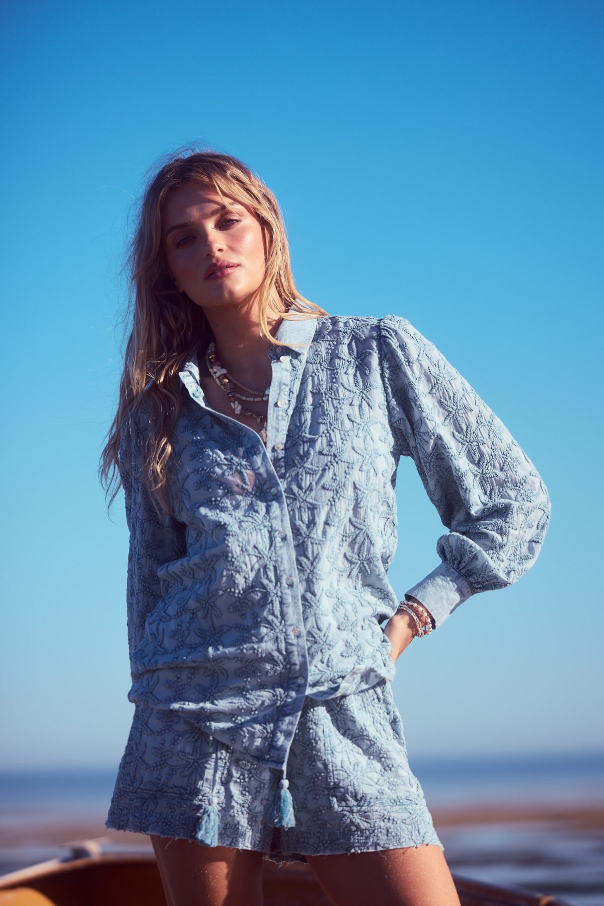 Flower Child Shirt in powder blue, standing by the shore under a blue sky.