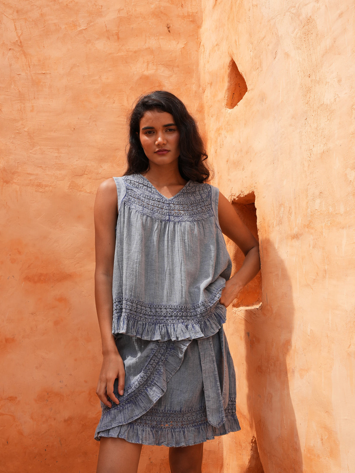 Freesia Mini Wrap Skirt and Freesia Top in steel blue, leaning on a terracotta wall.