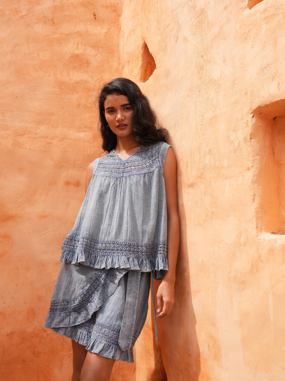 Freesia Top in steel blue with the Ithika Short, leaning on a terracotta wall.