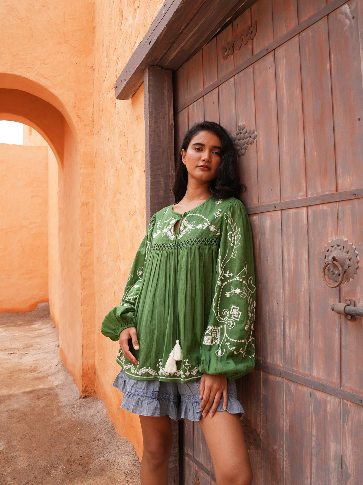 Sapphire Top in olive with the Ithika Short, standing at a weathered timber door.