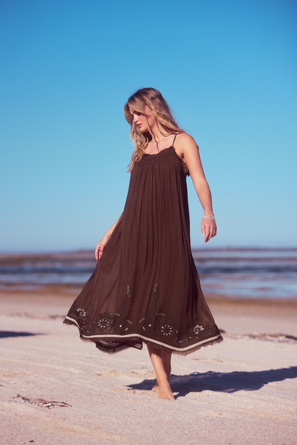 Ambrosia Maxi Dress in cocoa, mid-stride on the beach under a blue sky.