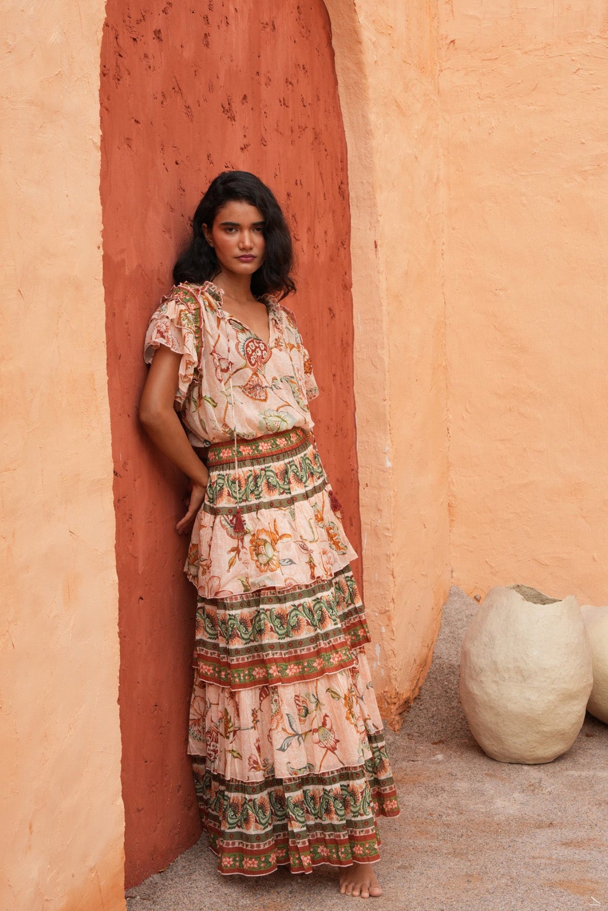 A woman stands barefoot in a rustic outdoor setting with textured orange walls, wearing a long tiered bohemian maxi dress with a floral and geometric print. Large clay pots sit on the ground to her right.
